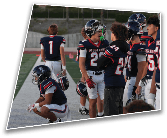 A group of people wearing football uniforms and helmets are standing and sitting on the sidelines of a football field, with one person in casual clothing among them. Description generated by AI