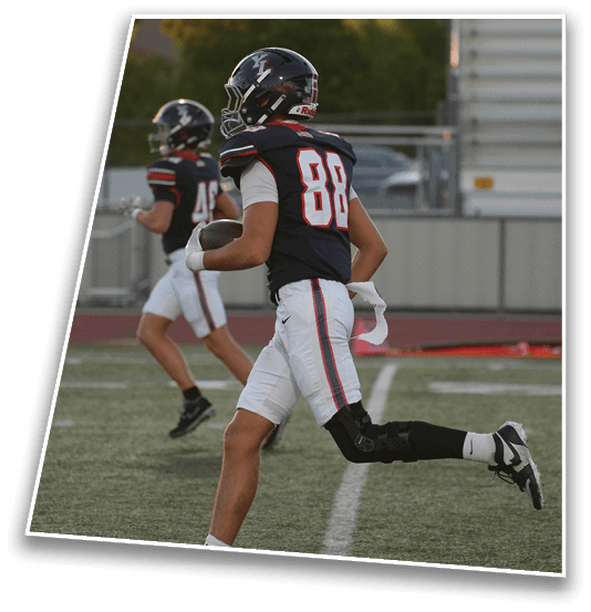 A person wearing a dark sports jersey with the number 88 and a helmet is standing outdoors near a fence with trees and a building in the background during sunset. Description generated by AI