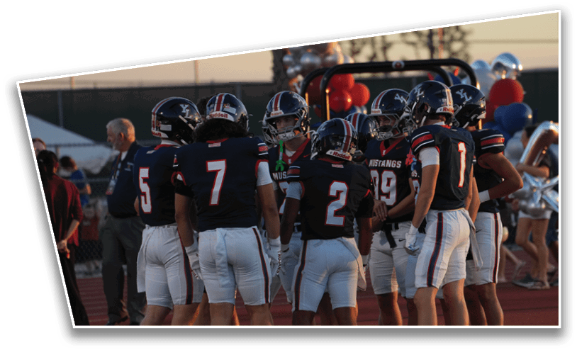 A group of people wearing football uniforms are standing together on a field with a football on the ground near them and balloons and cheerleaders visible in the background. Description generated by AI