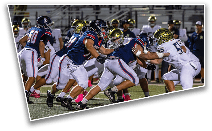 Players in football uniforms are engaged in a close, intense play on a field with a stadium background. Description generated by AI