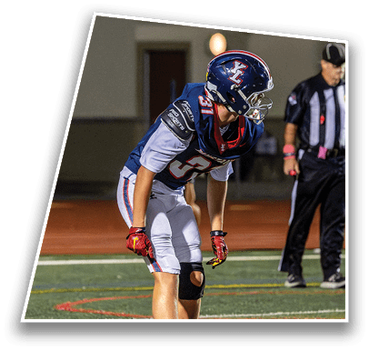 A person in a football uniform and helmet is bent forward in a ready stance on a field while a referee stands in the background. Description generated by AI