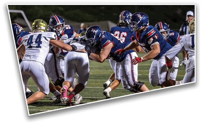 Players in football uniforms are engaged in a close, intense play on a well-lit field during a game. Description generated by AI