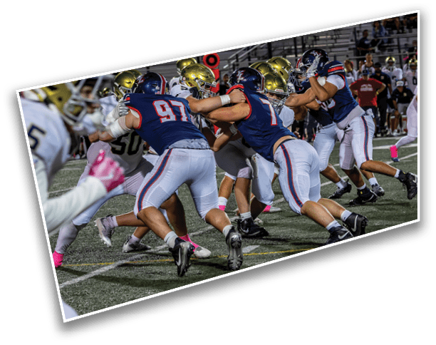 Players in football uniforms are engaged in a close, intense play on a well-lit field during a game. Description generated by AI