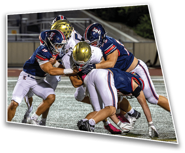 Several football players in helmets and uniforms are engaged in a close tackle on a field during a game. Description generated by AI