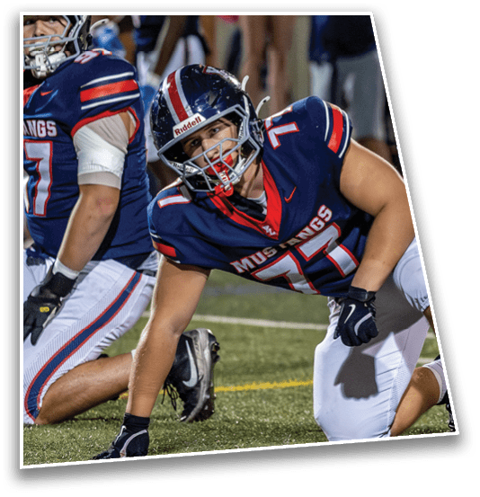 A person wearing a blue football uniform with the number 71 and the word Mustangs is crouched on a grassy field, preparing for a play. Description generated by AI