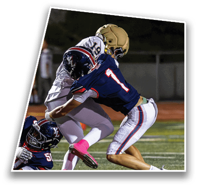 Two people in football uniforms are engaged in a tackle on a field during a game, with one player wearing a gold helmet and white uniform and the other wearing a navy blue uniform with red and white stripes. Description generated by AI