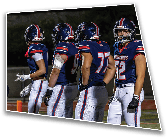 Four people in football uniforms and helmets stand side by side on a field at night, with one person facing forward and the others facing sideways. Description generated by AI