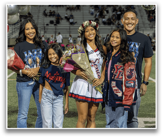 Four people stand on a sports field smiling, with one wearing a cheerleading outfit and sash while holding flowers and another holding a bouquet of flowers. Description generated by AI