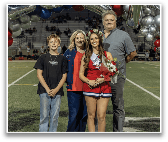 Four people stand together on a sports field with one person wearing a cheerleading outfit, a floral crown, and holding a bouquet of flowers, while the others stand close by smiling. Description generated by AI