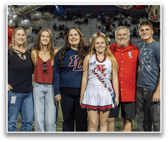 Five people stand together on a sports field with balloons in the background, including one person wearing a cheerleading uniform and a floral crown with a sash that reads “Senior." Description generated by AI