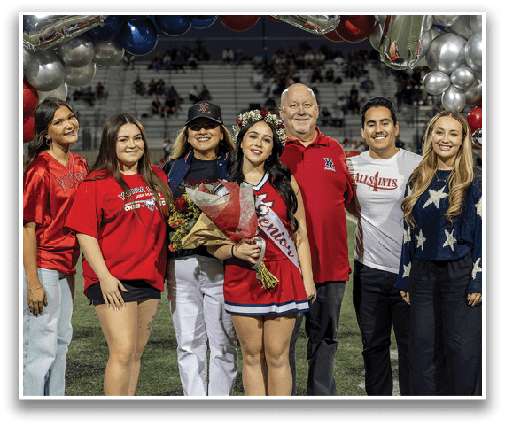 A group of six people stand on a sports field with one person wearing a sash and crown holding a bouquet of flowers, while others smile and wear casual and team-themed clothing. Description generated by AI