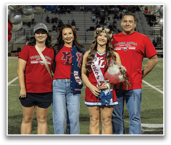 Four people stand on a sports field with balloons in the background, three wearing red shirts with “Yorba Linda High School Cheer Song" text, one wearing a cheerleader outfit with a sash and flower crown holding a bouquet. Description generated by AI