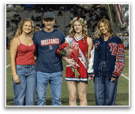 Four people stand on a sports field at night with one person wearing a cheerleader outfit, a flower crown, and a sash while holding flowers, and the others dressed casually. Description generated by AI