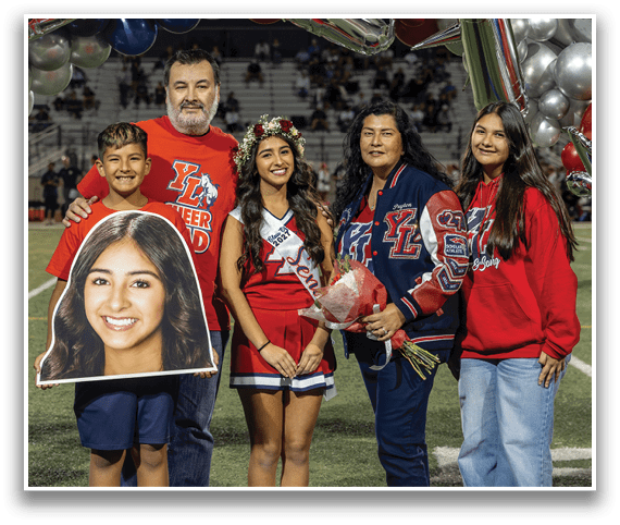 Four people stand on a sports field with balloons in the background, one person holding a large cutout of a smiling face, another wearing a cheerleader outfit and a flower crown, and another holding a bouquet of flowers. Description generated by AI