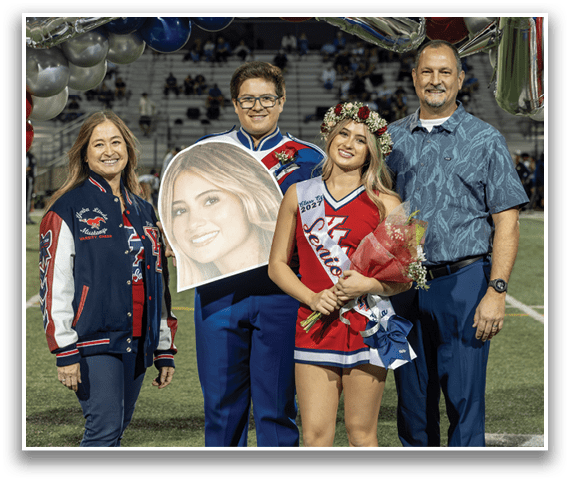 Four people stand on a sports field under a balloon arch, with one person holding a large cutout of a smiling face and another wearing a floral crown and a sash that reads “Senior." Description generated by AI