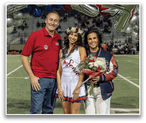 A person wearing a red shirt stands next to a person in a cheerleader outfit with a flower crown and sash, and another person holding a bouquet of flowers, all smiling on a sports field. Description generated by AI