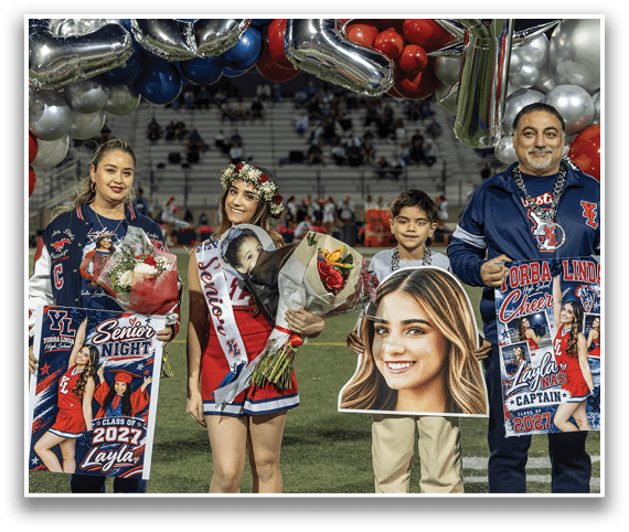 Three people stand on a sports field holding flowers, a banner, and large cutout photos, with one person wearing a floral crown and a sash that reads “Senior." Description generated by AI