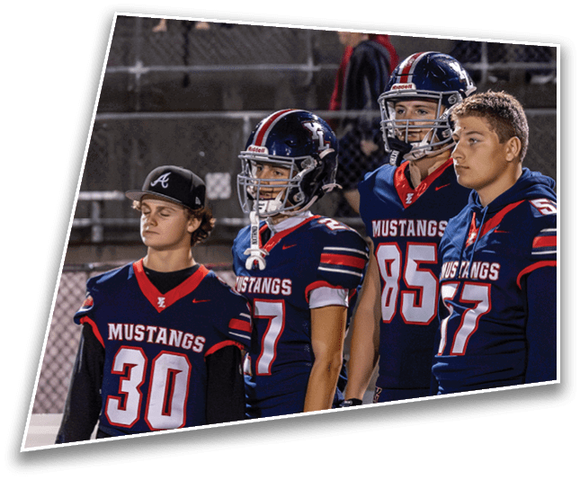 Four people wearing Mustangs football uniforms stand side by side on a sports field at night, with one person wearing a baseball cap and the others in helmets or sports gear. Description generated by AI