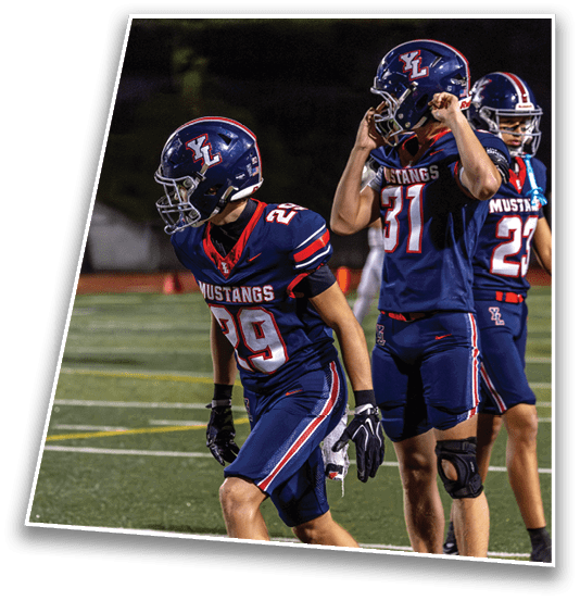 Two people wearing navy blue football uniforms with “Mustangs" on the front and helmets are standing on a football field, one looking forward and the other adjusting their helmet. Description generated by AI