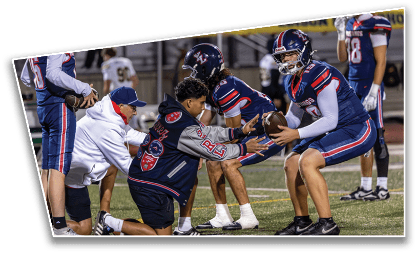 People in football uniforms and a coach in a white hoodie are on a field with one player handing a football to another during practice or a game. Description generated by AI