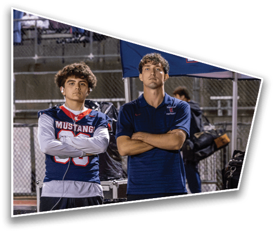 Two people with crossed arms stand side by side on a sports field, one wearing a football jersey with “Mustangs" and the other in a polo shirt. Description generated by AI