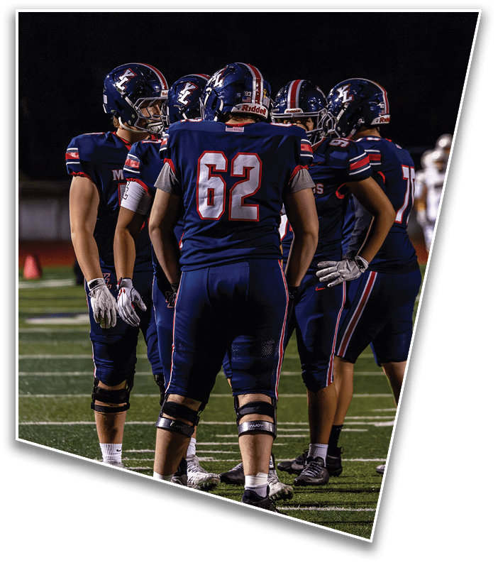Five people in football uniforms and helmets stand together on a field at night, appearing to discuss strategy. Description generated by AI