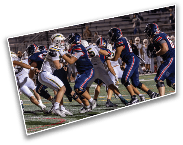 Players in dark and white football uniforms are engaged in a play on a field with empty bleachers in the background. Description generated by AI