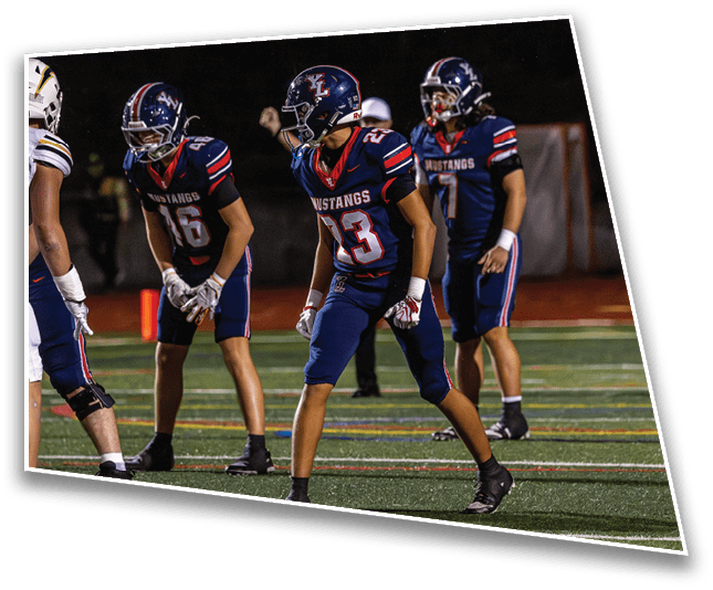 Three people in navy blue football uniforms with “Mustangs" on the jerseys are positioned on a football field at night, appearing ready for a play. Description generated by AI