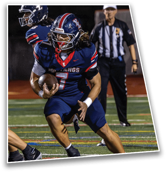 A person wearing a blue football uniform with the number seven is running with a football on a field while a referee stands in the background. Description generated by AI