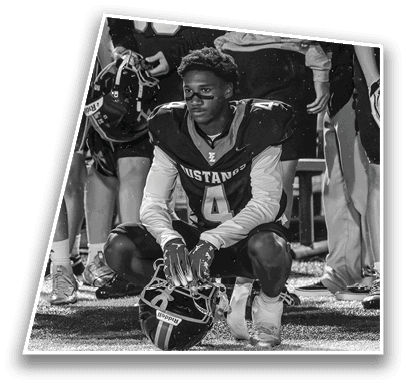 A person wearing a Mustangs football uniform with the number 4 is sitting on a bench looking forward while other players stand behind. Description generated by AI