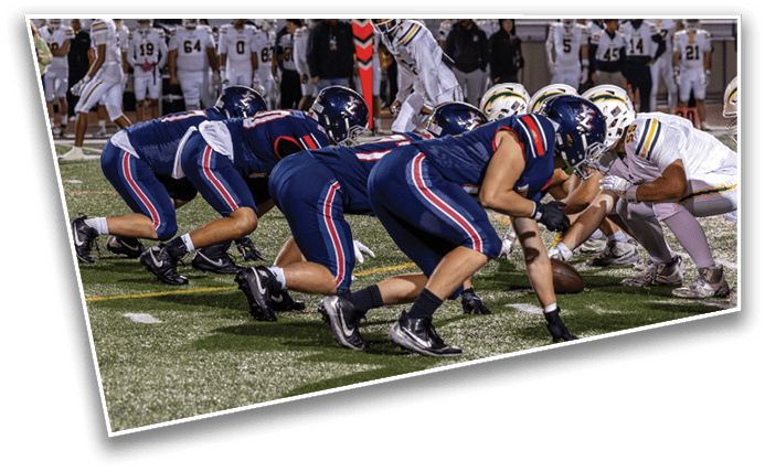 Two opposing American football teams are lined up at the line of scrimmage on a field with players in blue and white uniforms preparing for the next play. Description generated by AI