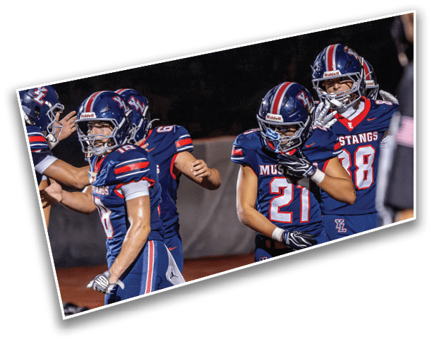 Four people wearing blue football uniforms with helmets are standing on a field, with two of them appearing to be in conversation or celebration. Description generated by AI