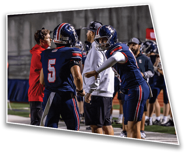 People wearing football helmets and uniforms are gathered on the sidelines during a nighttime game. Description generated by AI