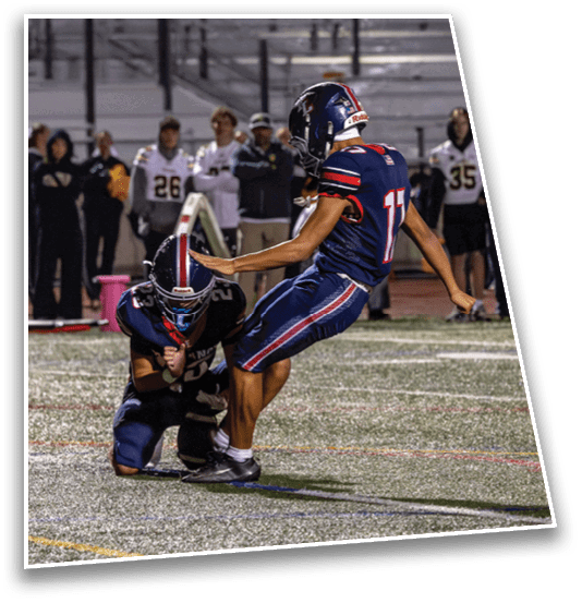 Two people in football uniforms are on a field with one holding the ball on the ground while the other prepares to kick it during a nighttime game. Description generated by AI