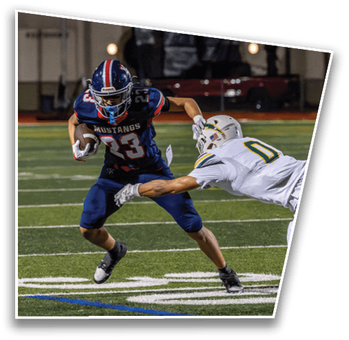 A person in a blue Mustangs football uniform is running with the ball while another person in a white football uniform attempts to tackle them on a lit football field. Description generated by AI
