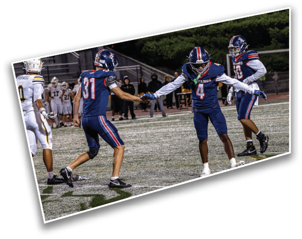 Three people in blue football uniforms are celebrating on a football field while other players and spectators are visible in the background. Description generated by AI