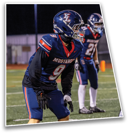 Two people in blue football uniforms with helmets are positioned on a field, appearing ready for play. Description generated by AI