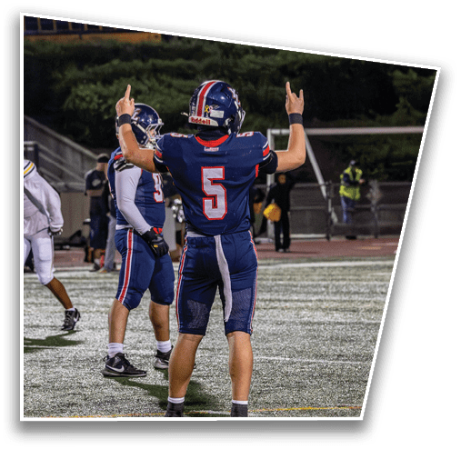 A person wearing a blue football uniform with the number 5 is raising both hands with index and pinky fingers extended on a football field at night while another person in a similar uniform stands nearby. Description generated by AI