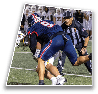 A person in a dark blue football uniform tackles another person in a white football uniform on a field while a referee closely watches and a group of people in white uniforms stand in the background near a yellow tent. Description generated by AI