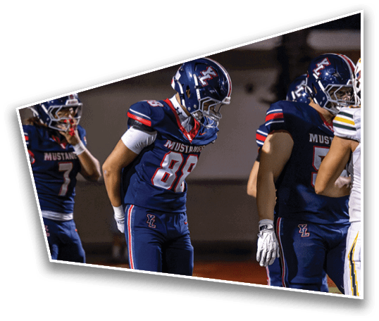 Three people in football uniforms with helmets stand on a field, with one person in the center looking down and the others standing nearby. Description generated by AI