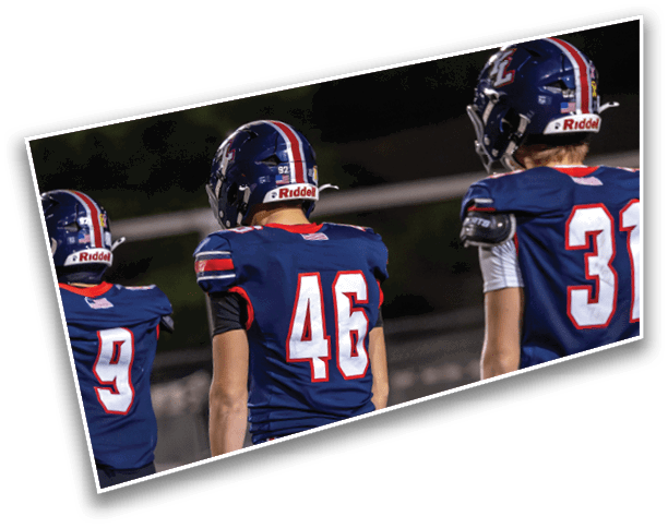 Three people wearing blue football uniforms with helmets stand side by side on a field at night. Description generated by AI