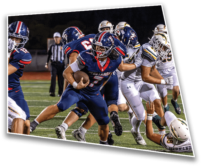 Several football players in blue and white uniforms are actively engaged in a game on a field, with one player in blue holding the ball and attempting to advance while others block or tackle. Description generated by AI