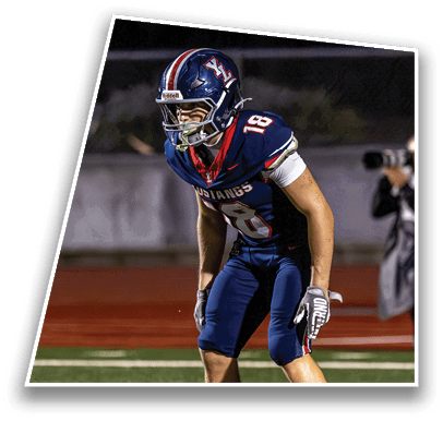 A person wearing a blue football uniform with the number 18 is crouched in a ready position on a football field. Description generated by AI