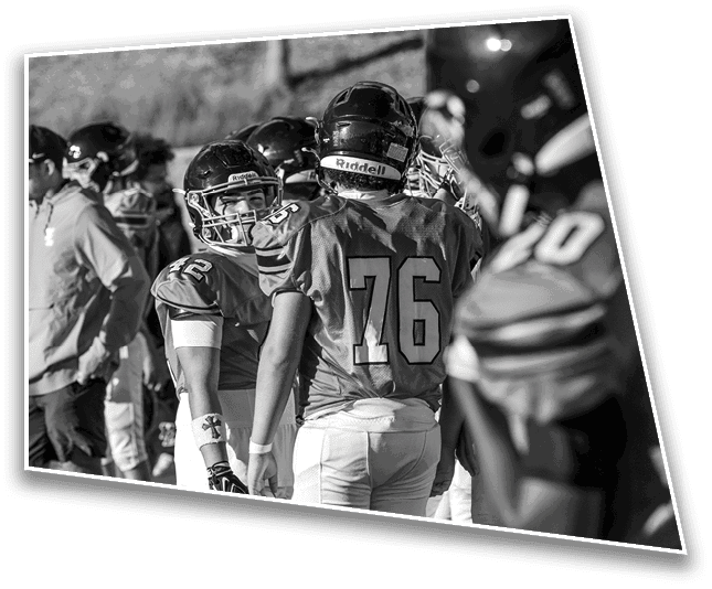 People wearing football helmets and uniforms are gathered closely together outdoors, seemingly preparing or strategizing during a game or practice. Description generated by AI