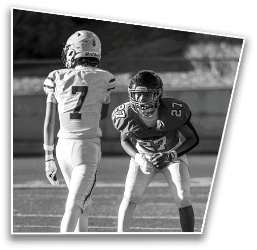 Two people wearing football uniforms and helmets are positioned on a field, one standing upright and the other crouched in a ready stance. Description generated by AI