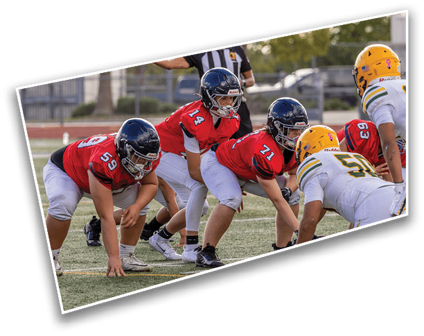People in football uniforms are lined up on a field preparing for a play, with one person in a red jersey positioned behind the line of scrimmage. Description generated by AI