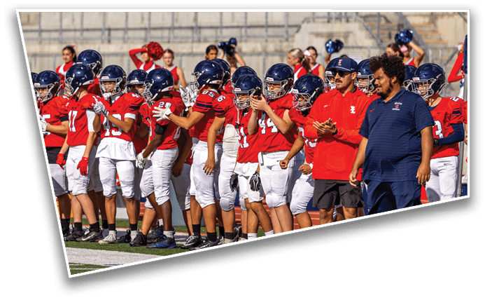 A group of people wearing red sports jerseys and helmets stand on a field with two adults in casual clothing standing nearby. Description generated by AI