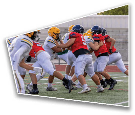 People wearing helmets and sports uniforms are engaged in a football game on a field with a track and a fence in the background. Description generated by AI
