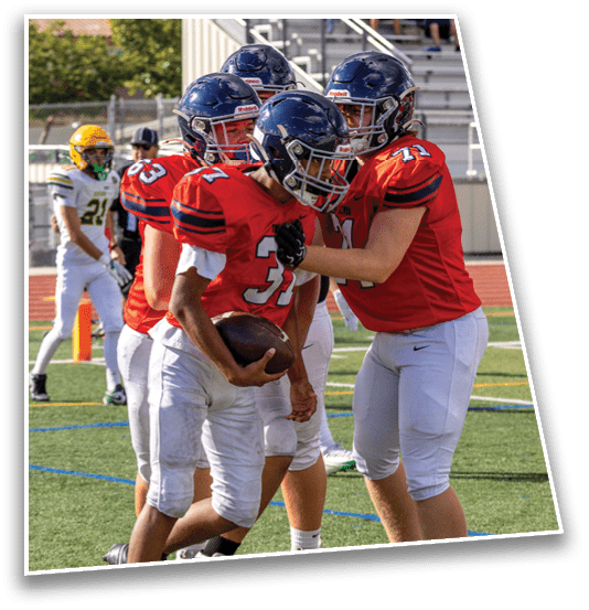 Four people in football uniforms are on a field with one holding a football while others appear to be celebrating or encouraging, and a person in a different uniform stands in the background near the sideline. Description generated by AI