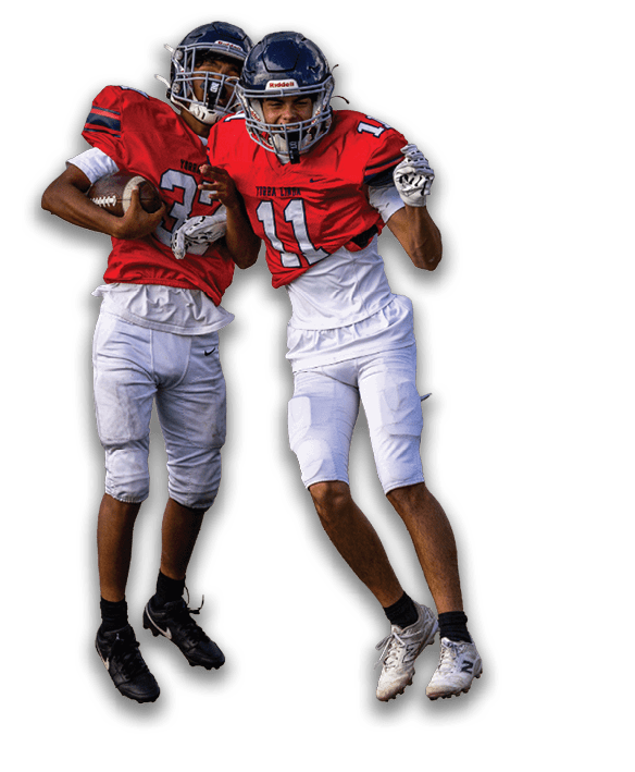 Two people wearing red football jerseys and white pants are jumping with one holding a football and the other pointing forward. Description generated by AI